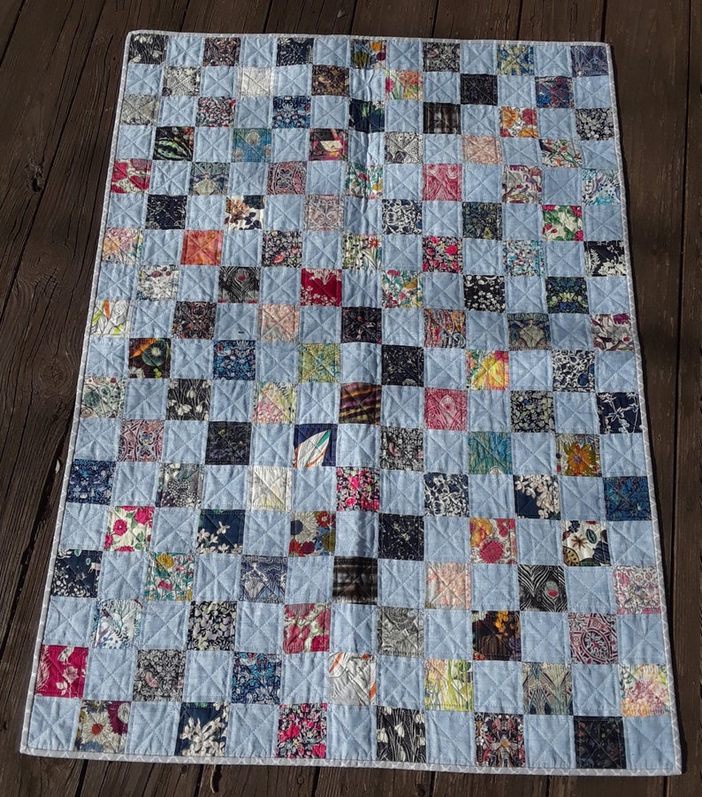 Pu&ograve; includere: Una trapunta patchwork con sfondo azzurro e una griglia di quadrati colorati. I quadrati presentano tessuti floreali e fantasia in vari colori, tra cui rosa, blu e verde. La trapunta misura circa 91 cm di larghezza e 122 cm di lunghezza.