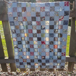 Pu&ograve; includere: Una trapunta patchwork fatta a mano con uno sfondo azzurro e un motivo a griglia di quadrati colorati. I quadrati presentano vari tessuti floreali e fantasia nei toni del rosso, blu e marrone. La trapunta &egrave; appesa a una staccionata di legno.