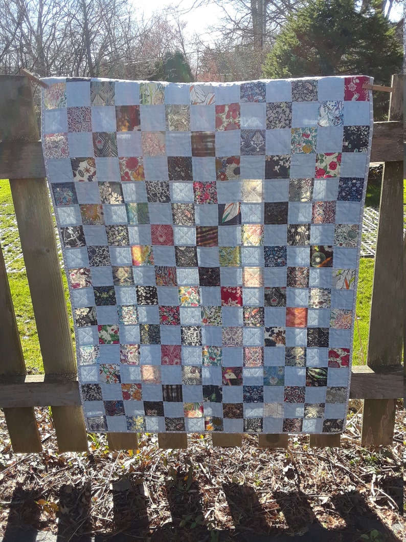 Pu&ograve; includere: Una trapunta patchwork fatta a mano con uno sfondo azzurro e un motivo a griglia di quadrati colorati. I quadrati presentano vari tessuti floreali e fantasia nei toni del rosso, blu e marrone. La trapunta &egrave; appesa a una staccionata di legno.