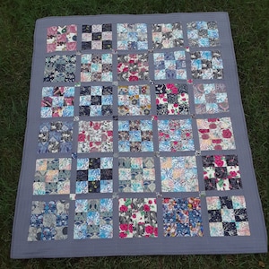 Può includere: Una trapunta grigia con un design a patchwork. La trapunta è composta da molti quadrati di tessuto diversi, ognuno con un motivo unico. La trapunta è un bellissimo esempio di artigianato fatto a mano.