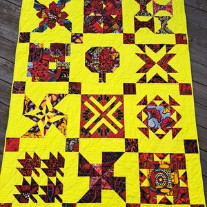 May include: A yellow quilt with a patchwork design featuring red, black, and white fabric squares. The quilt is made up of various geometric patterns, including stars, diamonds, and triangles.