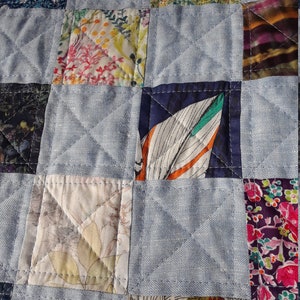 Pu&ograve; includere: Primo piano di una trapunta patchwork con sfondo azzurro e toppe quadrate di vari tessuti floreali e fantasia. La trapunta &egrave; cucita con un motivo a rombi, che mostra la lavorazione e il design dettagliati.