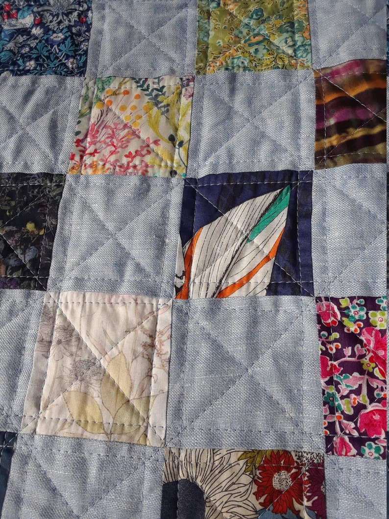 Pu&ograve; includere: Primo piano di una trapunta patchwork con sfondo azzurro e toppe quadrate di vari tessuti floreali e fantasia. La trapunta &egrave; cucita con un motivo a rombi, che mostra la lavorazione e il design dettagliati.