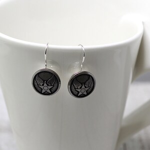 May include: A pair of silver dangle earrings with a circular design featuring a star and wings. The earrings are hanging from a white mug.