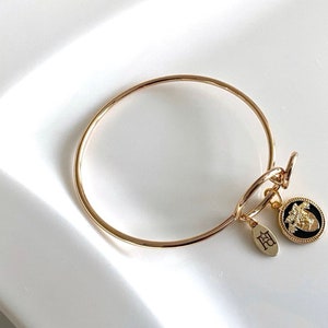 May include: A gold bangle bracelet with a black and gold charm. The charm features a floral design and the letters "FB".