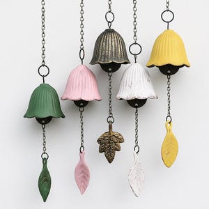 May include: A collection of six hanging wind chimes. Each chime features a bell-shaped top in various colors: green, pink, bronze, white, and yellow. Each chime has a leaf-shaped or leaf-like pendant. The chimes are suspended by chains.