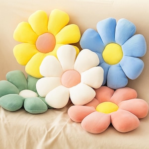 May include: Five flower-shaped pillows in various colors: yellow, blue, white, green, and pink. Each pillow has a contrasting center, with a ribbed texture. The pillows are arranged on a beige surface.