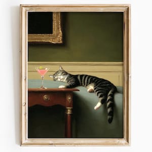 Op de afbeelding: Een ingelijste kunstdruk met een ontspannen tijgerkat naast een roze cocktail. De kat ligt op een turquoise oppervlak, met een kleine, donkere houten tafel op de voorgrond. De achtergrond is een donkergroene muur met een goudomrand schilderij.