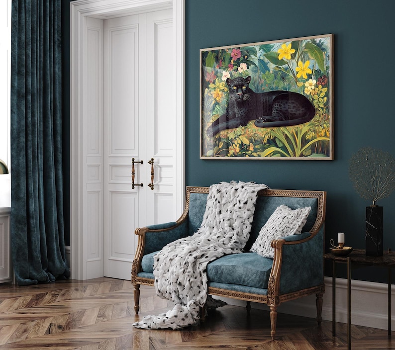 Op de afbeelding: Een ingelijste kunstdruk met een zwarte panter die rust in een weelderige, kleurrijke junglesc&egrave;ne met gele en roze bloemen. Het kunstwerk hangt boven een blauwgroene bank met een decoratief kleed en kussen.