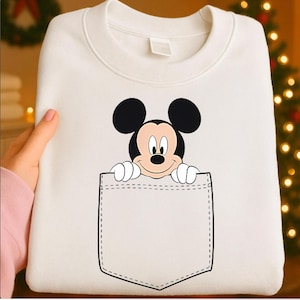 May include: White sweatshirt featuring a graphic of Mickey Mouse peeking out of a pocket. The Mickey Mouse graphic is in black, red, and white. The pocket is outlined in black stitching.