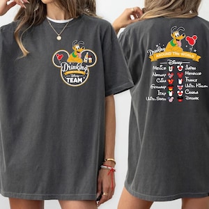 Könnte beinhalten: Ein anthrazitfarbenes T-Shirt mit einem Brustmotiv von Pluto in einer Micky-Maus-Ohren-Silhouette und dem Text Drinking Around The World Disney Team. Auf der Rückseite ist Pluto sowie eine Liste von Ländern mit Flaggen-Symbolen abgebildet.