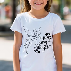 Puede incluir: Camiseta blanca con una ilustración negra de un hada y el texto "Think Happy Thoughts" y estrellas. La camiseta tiene mangas cortas y cuello redondo.