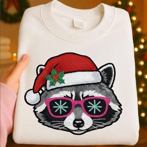 May include: White sweatshirt with an embroidered raccoon wearing a Santa hat and pink sunglasses with snowflake designs. The raccoon has gray and black fur. The Santa hat is red with a white trim and a sprig of holly.