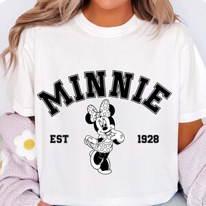 May include: White t-shirt featuring a black and white graphic of Minnie Mouse. The word "MINNIE" is arched across the top, with "EST 1928" below. Minnie Mouse is depicted in her classic polka dot bow and dress.