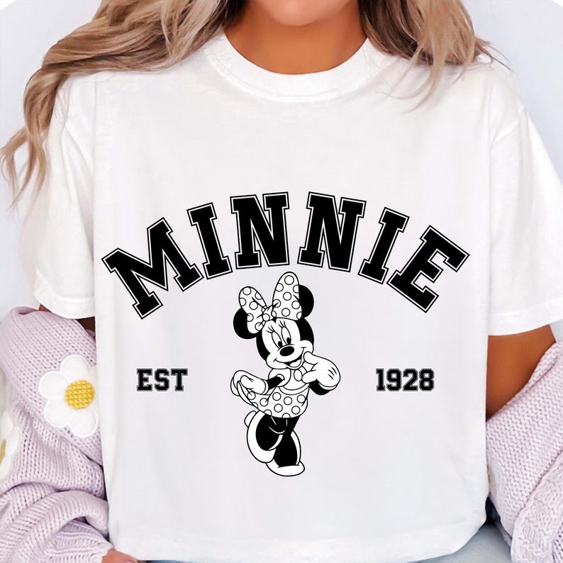May include: White t-shirt featuring a black and white graphic of Minnie Mouse. The word "MINNIE" is arched across the top, with "EST 1928" below. Minnie Mouse is depicted in her classic polka dot bow and dress.