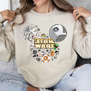 May include: A beige sweatshirt with a white Mickey Mouse silhouette featuring Star Wars characters, including Darth Vader, Yoda, R2-D2, and a TIE fighter. The text "STAR WARS" is printed in gold letters.