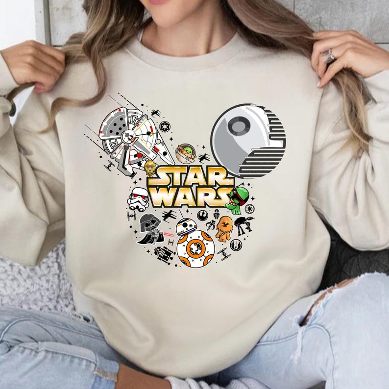 May include: A beige sweatshirt with a white Mickey Mouse silhouette featuring Star Wars characters, including Darth Vader, Yoda, R2-D2, and a TIE fighter. The text "STAR WARS" is printed in gold letters.