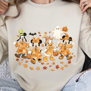 May include: Beige sweatshirt featuring a Halloween-themed graphic. The design includes cartoon characters Goofy, Minnie Mouse, Donald Duck, and Mickey Mouse, dressed in patchwork costumes, holding pumpkin-shaped treat buckets, and surrounded by autumn leaves and a ghost.