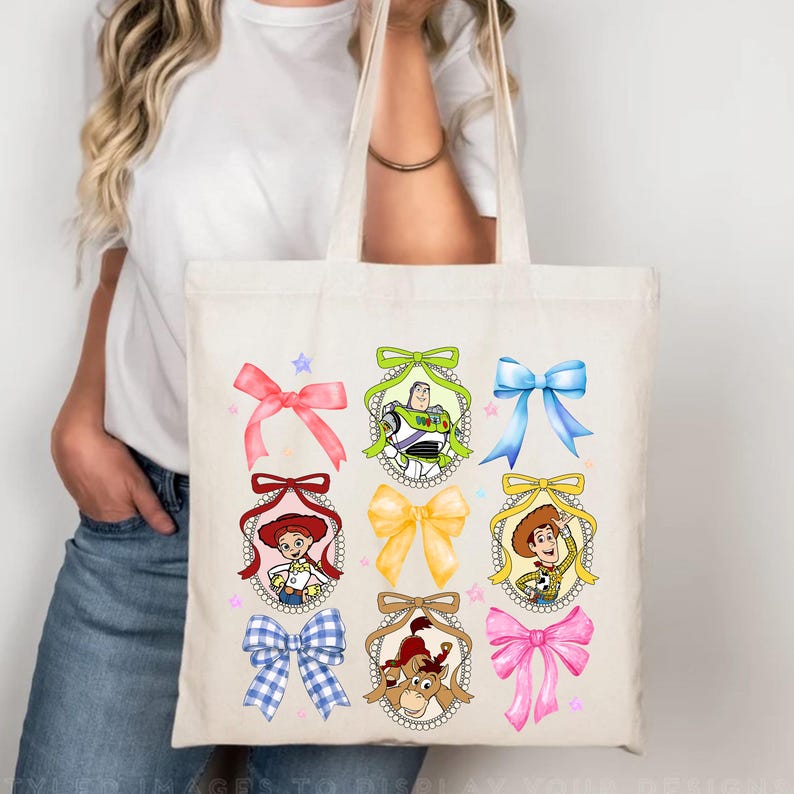 Puede incluir: Bolso tote crema con lazos coloridos y retratos de personajes de dibujos animados. El dise&ntilde;o incluye un lazo rosa, verde y azul, junto con retratos de Woody, Buzz Lightyear y Jessie. El bolso lo sostiene una persona.