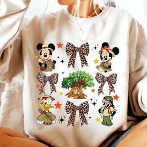 May include: Cream-colored sweatshirt featuring cartoon characters Mickey Mouse, Minnie Mouse, Donald Duck, and Goofy in safari outfits. The design includes leopard print bows, stars, and a tree graphic.