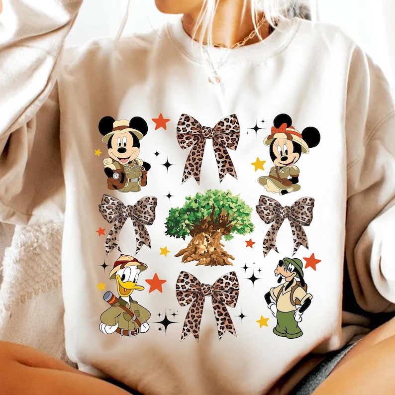 May include: Cream-colored sweatshirt featuring cartoon characters Mickey Mouse, Minnie Mouse, Donald Duck, and Goofy in safari outfits. The design includes leopard print bows, stars, and a tree graphic.