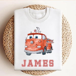 May include: White sweatshirt featuring a cartoon fire truck in shades of red and orange, with blue eyes and a smiling face. The name "JAMES" is printed in red block letters below the truck. A fun, playful design.