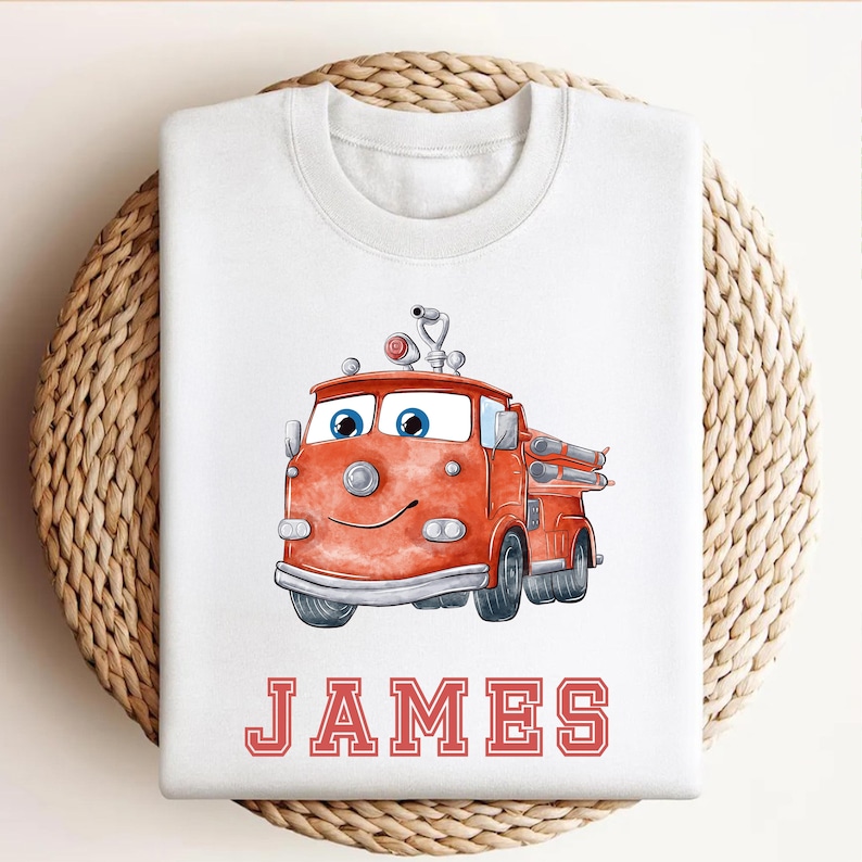 May include: White sweatshirt featuring a cartoon fire truck in shades of red and orange, with blue eyes and a smiling face. The name "JAMES" is printed in red block letters below the truck. A fun, playful design.