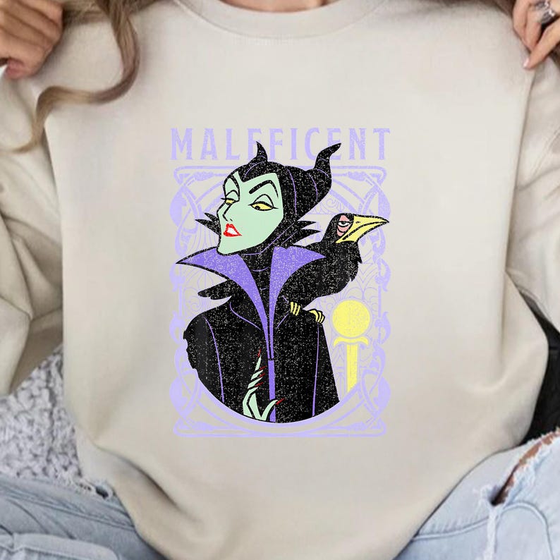 May include: A light beige sweatshirt featuring a graphic of Maleficent with her raven. The design includes the word "MALEFICENT" in lavender, with a green-skinned Maleficent and a black raven. The artwork is framed in a lavender decorative border.