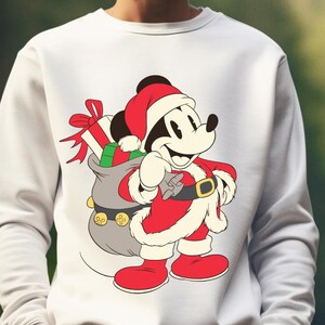 以下が含まれることがあります： クリスマスをテーマにしたミッキーマウスのデザインが施された白いスウェットシャツ。ミッキーはサンタクロースの格好をしており、赤いスーツと帽子を身に着け、プレゼントでいっぱいの灰色の袋を持っています。デザインはカラフルなイラストです。