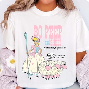 May include: White t-shirt with a cartoon graphic of Bo Peep and sheep. The design includes Bo Peep and three sheep, with the text "Bo Peep and Sheep Porcelain Figure Set." The text "ONLY! AT SELECT TOY STORES!" is also included in the design.