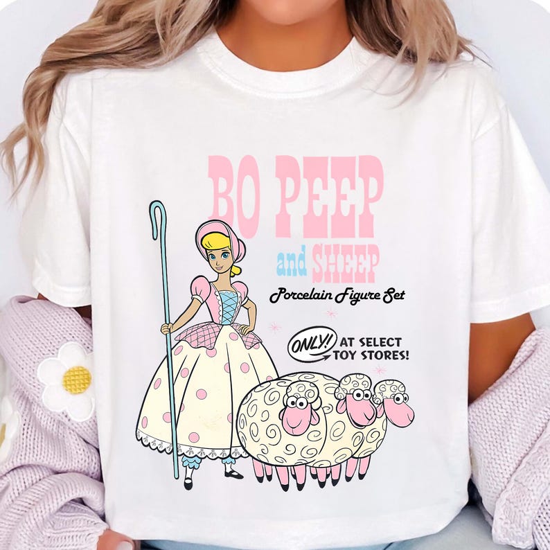 May include: White t-shirt with a cartoon graphic of Bo Peep and sheep. The design includes Bo Peep and three sheep, with the text "Bo Peep and Sheep Porcelain Figure Set." The text "ONLY! AT SELECT TOY STORES!" is also included in the design.