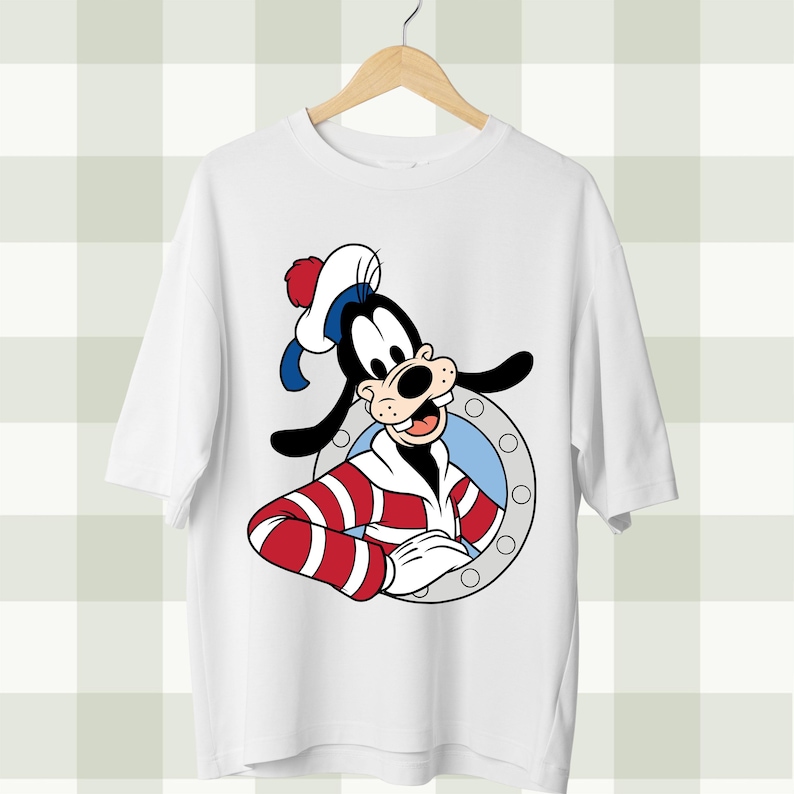May include: White t-shirt featuring a cartoon image of Goofy in a sailor outfit. Goofy is wearing a sailor hat and a red and white striped shirt. The design is set inside a circular frame.
