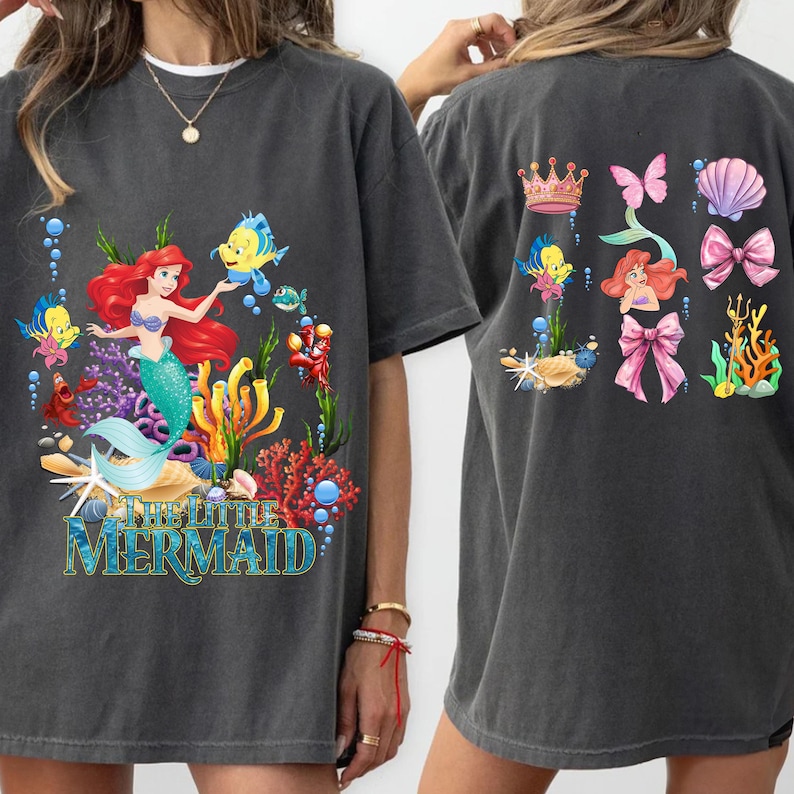 Op de afbeelding: Donkergrijs T-shirt met een kleurrijke afbeelding van Ariel uit De Kleine Zeemeermin, omringd door zeedieren en koraal. De achterkant toont extra zeemeermin-thema illustraties, waaronder een kroon, zeeschelp en strikken. De tekst "THE LITTLE MERMAID" staat onderaan.