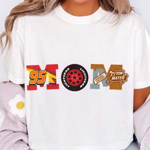 May include: White t-shirt with a colorful graphic design. The design spells "MOM" using elements from the movie Cars. The "M" features the number 95, the "O" is a red tire, and the second "M" includes Tow Mater.