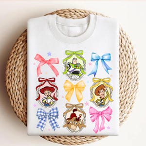 Puede incluir: Sudadera blanca con retratos de personajes de dibujos animados en marcos decorativos, cada uno adornado con un lazo colorido. El dise&ntilde;o incluye personajes como Buzz Lightyear y Woody, con estrellas rosas y azules.