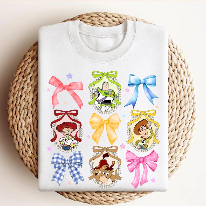 Puede incluir: Sudadera blanca con retratos de personajes de dibujos animados en marcos decorativos, cada uno adornado con un lazo colorido. El dise&ntilde;o incluye personajes como Buzz Lightyear y Woody, con estrellas rosas y azules.