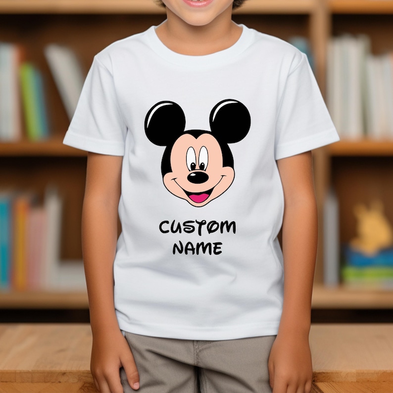 May include: White t-shirt featuring a large graphic of Mickey Mouse's face in black, red, and beige. The text "CUSTOM NAME" is printed in black below the graphic. The shirt is short-sleeved.