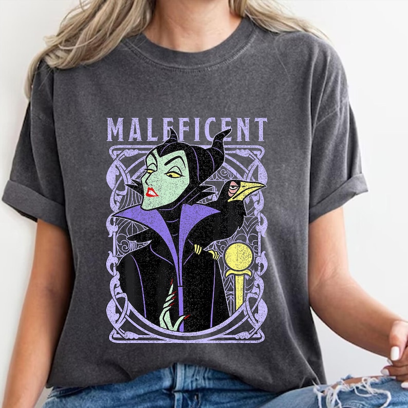 May include: Dark gray t-shirt featuring a graphic of Maleficent with her raven. The design includes the word "Maleficent" in a lavender font. The artwork is framed by a decorative lavender border. The shirt is a casual wear item.