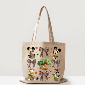 May include: Beige tote bag featuring Mickey Mouse, Minnie Mouse, Donald Duck, and Goofy in safari outfits. The bag also has leopard print bows, a tree graphic, and star accents. A fun and whimsical design.
