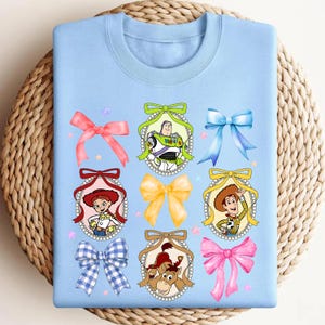 Puede incluir: Sudadera azul claro con personajes de dibujos animados y lazos coloridos. El dise&ntilde;o incluye im&aacute;genes de Buzz Lightyear, Jessie, Woody y Bullseye, cada uno enmarcado con un borde decorativo. Tambi&eacute;n se muestran varios lazos en tonos rosa, amarillo y azul.