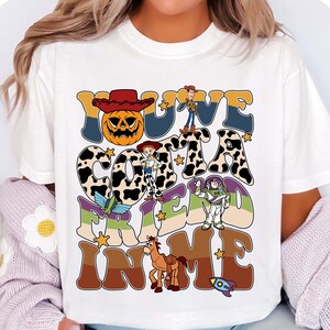 May include: White t-shirt with a colorful graphic design featuring the text "YOU'VE GOT A FRIEND IN ME" with cartoon characters and a pumpkin. The design includes a cowboy hat, stars, and a rocket.