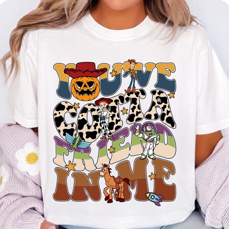 May include: White t-shirt with a colorful graphic design featuring the text "YOU'VE GOT A FRIEND IN ME" with cartoon characters and a pumpkin. The design includes a cowboy hat, stars, and a rocket.