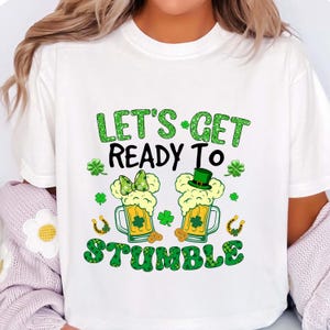 May include: White t-shirt with a St. Patrick's Day graphic. The design features two mugs of beer with foam, shamrocks, and the text "LET'S GET READY TO STUMBLE" in green and black. Includes a green top hat and bow.