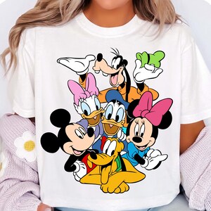 K&ouml;nnte beinhalten: Wei&szlig;es T-Shirt mit einem farbenfrohen Aufdruck von Disney-Figuren, darunter Mickey Mouse, Minnie Mouse, Donald Duck, Daisy Duck, Goofy und Pluto. Die Zeichentrickfiguren l&auml;cheln. Das Shirt ist aus weichem Material.