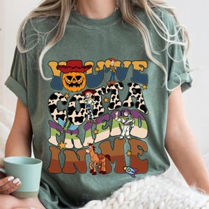 May include: Sage green t-shirt with a colorful graphic design. The design features the text "You've Gotta Friend In Me" with cartoon characters and a pumpkin wearing a cowboy hat. The shirt has short sleeves and a crew neck.