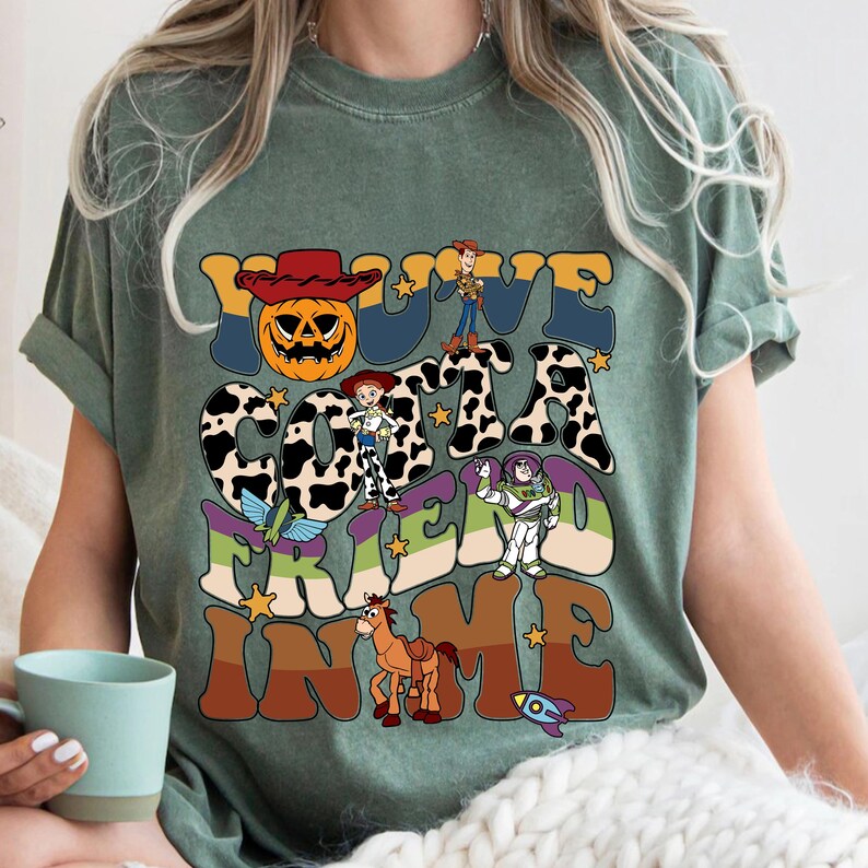 May include: Sage green t-shirt with a colorful graphic design. The design features the text "You've Gotta Friend In Me" with cartoon characters and a pumpkin wearing a cowboy hat. The shirt has short sleeves and a crew neck.