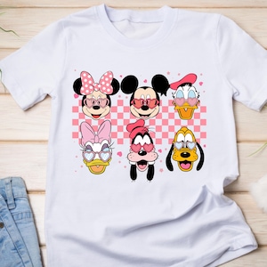 K&ouml;nnte beinhalten: Wei&szlig;es T-Shirt mit einem farbenfrohen Aufdruck von Disney-Figuren, darunter Minnie und Mickey Mouse, Donald Duck, Daisy Duck, Goofy und Pluto, alle mit herzf&ouml;rmigen Sonnenbrillen. Der Hintergrund hat ein rosa-wei&szlig;es Karomuster.