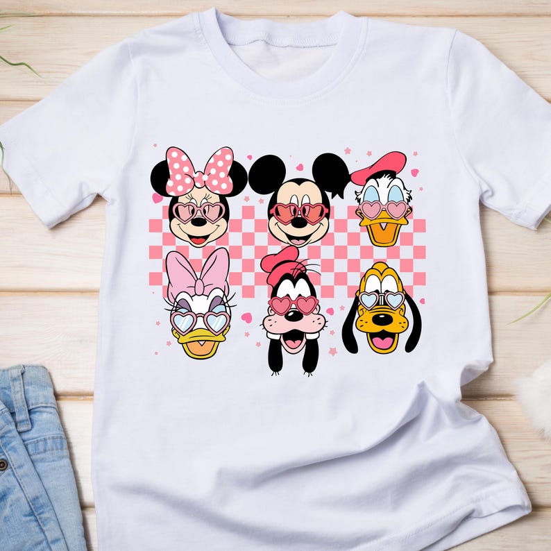 K&ouml;nnte beinhalten: Wei&szlig;es T-Shirt mit einem farbenfrohen Aufdruck von Disney-Figuren, darunter Minnie und Mickey Mouse, Donald Duck, Daisy Duck, Goofy und Pluto, alle mit herzf&ouml;rmigen Sonnenbrillen. Der Hintergrund hat ein rosa-wei&szlig;es Karomuster.