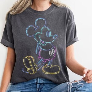 May include: Dark gray t-shirt featuring a neon-style Mickey Mouse design. The graphic uses vibrant colors, including blue, yellow, pink, and purple, to outline the iconic character. The shirt has short sleeves and a relaxed fit.