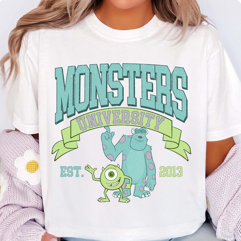 Puede incluir: Camiseta blanca con un gr&aacute;fico verde y azul de los personajes de la Universidad de Monstruos. El gr&aacute;fico incluye el texto "Monsters University" y "Est. 2013".